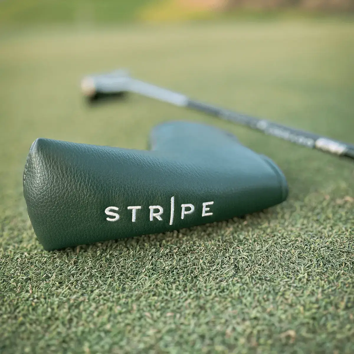 A green leather headcover on the putting green with a golf club, featuring the Stripe Golf logo in white embroidery.